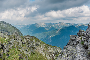 Close-up view of a rocky range behind which reveals panoramic view of an impressive mountain with high peaks illuminated by the sun and dramatic  sky covered with clouds.