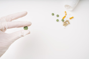 Hand in medicine disposable glove with green pill on the forefront. Medical pills and capsules spilled out from white plastic bottle on the background. Selective focus.