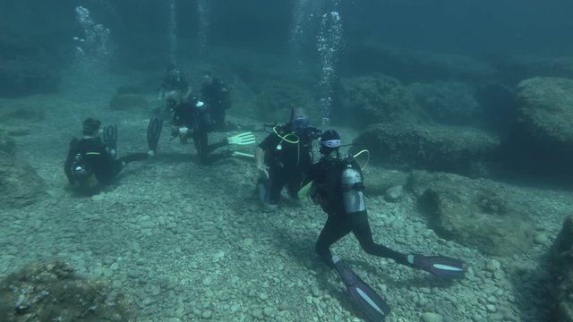 A diving instructor conducts lessons with beginner scuba divers on pebbly seabed among rocky reefs. An master dive trainer provides training for group of novice underwater explorers.