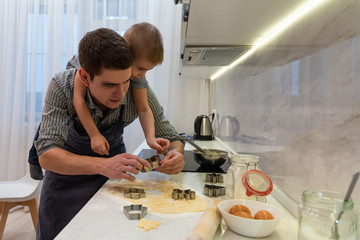Dad and son are preparing cookies in the kitchen. The boy hangs on the back of his parent and reaches for cookie cutters with his hand. Fun family tradition to cook together on World Womens Day.