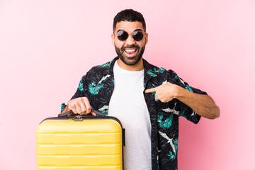 Young latin man holding a suitcase surprised pointing at himself, smiling broadly.