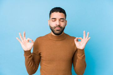 Fototapeta premium Young latin man against a blue background isolated relaxes after hard working day, she is performing yoga.