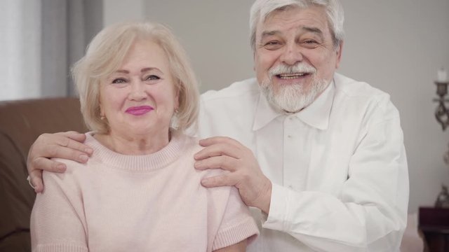 Elderly Caucasian couple talking to each other, looking at camera and smiling. Positive married retirees posing indoors. Love, happiness, lifestyle.