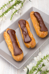 Tasty eclairs on a plate on a white wooden table. Served with flowers. Rectangular gray plate. Traditional french eclairs with chocolate. Home made cake eclairs. Traditional eclairs, profitroles class