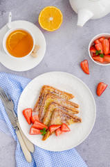 Traditional French toast sprinkled with powdered sugar with fresh strawberries on a white plate with a cup of tea. Delicious breakfast. Vertical orientation. Top view.