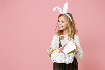 Obraz na płótnie Canvas Little pretty blonde kid girl 11-12 years old in spring dress, bunny rabbit ears on head hold in hand wicker basket colorful eggs gift coupon isolated on pastel pink background. Happy Easter concept.