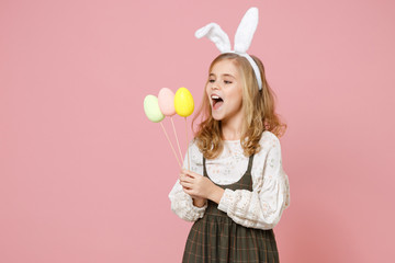 Obraz na płótnie Canvas Little pretty blonde kid girl 11-12 years old in spring dress, bunny rabbit ears hold in hand carries dyed eggs on sticks have fun celebrate isolated on pastel pink background. Happy Easter concept.