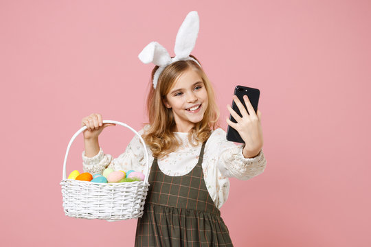 Little blonde kid girl 11-12 years old in spring dress bunny rabbit ears hold in hand cell phone wicker basket colorful eggs isolated on pastel pink background Childhood lifestyle Happy Easter concept