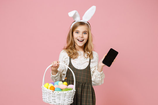 Little blonde kid girl 11-12 years old in spring dress bunny rabbit ears hold in hand cell phone wicker basket colorful eggs isolated on pastel pink background Childhood lifestyle Happy Easter concept