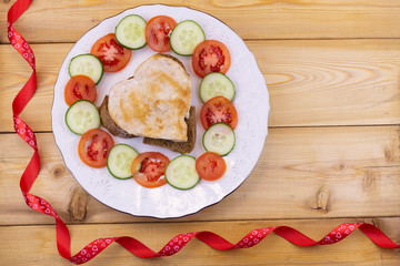 heart-shaped slice of chicken with cucumbers and tomatoes on a white plate. View from above. Wood background. Copy space