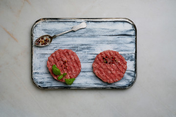 Raw Ground beef meat Burger steak cutlets with seasonings and herbs served on rustic wooden board with metal spoon over white marble background. Top view