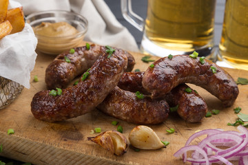 Grilled sausages from a BBQ served with a mustard, fried potatoes, garlic, herbs, beer on a wooden board all served on a black wooden table. Delicious german sausages Perfect for Octoberfest.