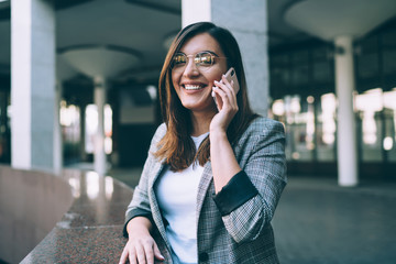 Pretty young woman in trendy eyewear look in camera enjoying mobile phone conversation on free time, happy female in spectacles laughing talking on smartphone via 4G internet connection