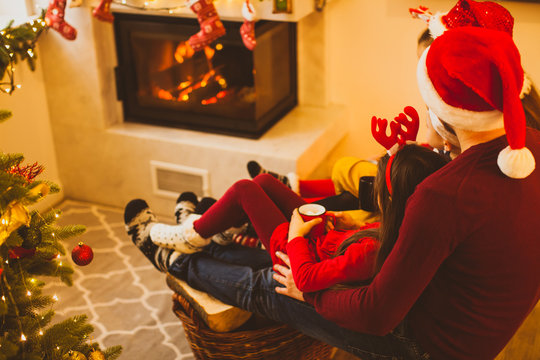 Cheerful family in Santa hats drinking tea near fireplace