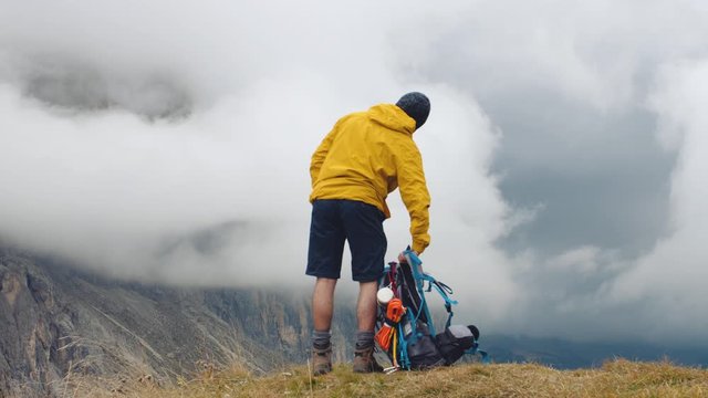 Young man hiker standing on mountain viewpoint with cloudy sky and fog. Yellow jacket, backpack, black beard and beanie. Traveling Dolomites, Italy.