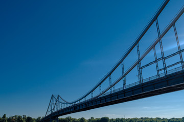 View of the long beautiful bridge from the side below. A pathway to the other side with trees against a blue sky without clouds on a clear day. Minimalist concept of joining with a place for text.