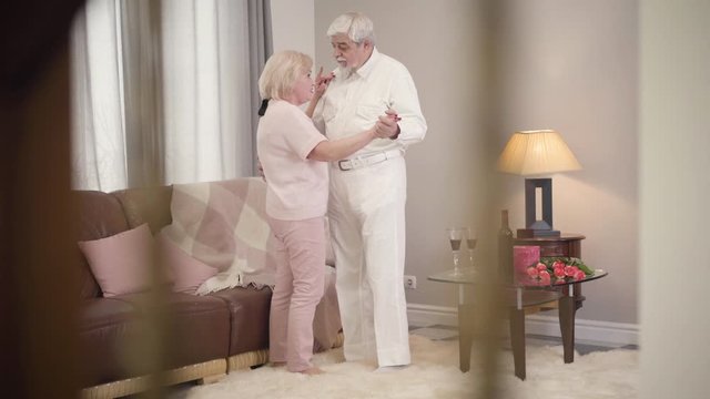 Long shot of mature Caucasian married couple talking and dancing at home. Happy senior retirees celebrating Saint Valentine's Day indoors. Eternal love of elegant spouses. Unity, bonding, lifestyle.