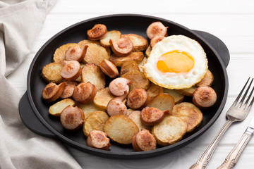 Close-up shot of fried potatoes with sausages and egg. Breakfast. Potatoes with sausages, cut into slices and fried in oil, in rustic style. Frying pan with tasty cooked egg and sausages on table