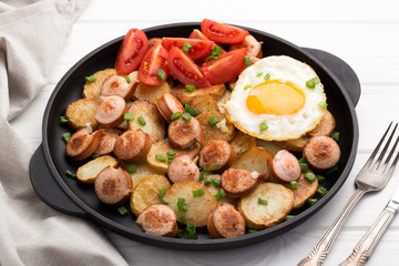 Close-up shot of fried potatoes with sausages and egg. Breakfast. Potatoes with sausages, cut into slices and fried in oil, in rustic style. Frying pan with tasty cooked egg and sausages on table