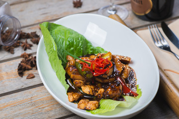 Sliced chicken fried in sweet and sour soy sauce with vegetables and herbs in a deep plate on a wooden table in a restaurant.