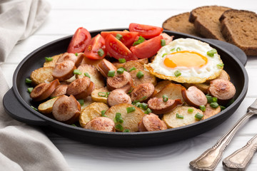 Fototapeta premium Close-up shot of fried potatoes with sausages and egg. Breakfast. Potatoes with sausages, cut into slices and fried in oil, in rustic style. Frying pan with tasty cooked egg and sausages on table