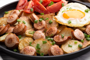Close-up shot of fried potatoes with sausages and egg. Breakfast. Potatoes with sausages, cut into slices and fried in oil, in rustic style. Frying pan with tasty cooked egg and sausages on table