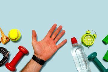 Flat lay shoot of green alarm clock, tennis ball, bottle of water, measuring tape, male hand and dumbbells on pastel blue backround. Top view with copy space for text. Fitness concept.