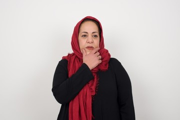 Isolated portrait of stylish muslim woman with hand under chin and looking sideways with doubtful and skeptical expression, worry and doubt. Standing indoors over gray background.