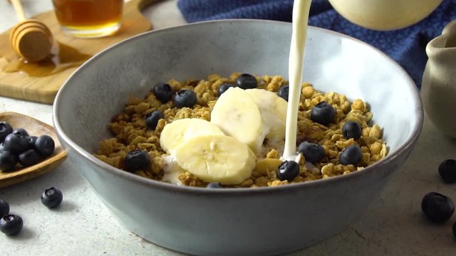 Healthy granola breakfast consisting of cereal, blueberries, banana and honey. Milk is poured in slow motion.