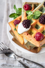 A stack of homemade Belgian waffles with frozen berries, mint and natural greek yogurt on a white plate, light grey stone background. Top view.