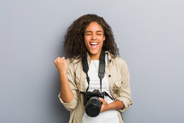 Young african american photographer woman holding a camera cheering carefree and excited. Victory concept. - Powered by Adobe