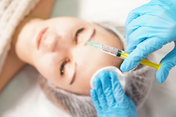 Rejuvenating facial injection treatment. Beautician holds an injection syringe. Woman face in the background. Focus on hand with syringe.
