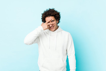 Young sporty man against a blue background blink at the camera through fingers, embarrassed covering face.