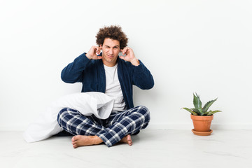 Mature man wearing pajama sitting on house floor covering ears with hands.