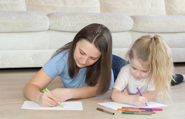 Young mother drawing coloring picture with cute daughter laying on warm floor