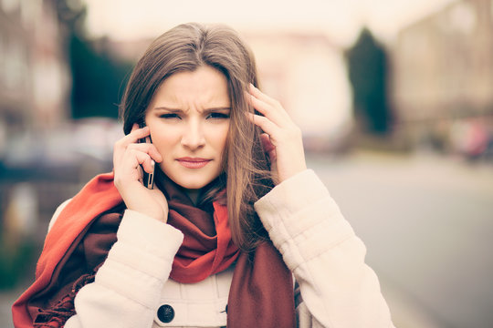 Phone talk. Portrait frustrated beautiful woman speaking on the mobile phone in a city street unfocused background having headache head pain problem stress depression hand on ear looking at you camera