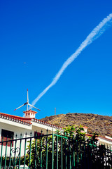 Estela de un avión que sale de las aspas de un aerogenerador con casa de campo en primer plano y cielo azul