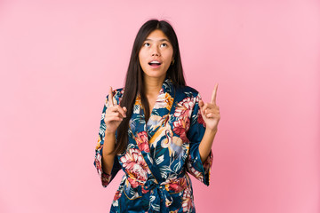 Young asian woman wearing a kimono pajamas pointing upside with opened mouth.