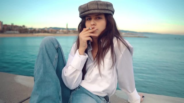 Portrait of young beautiful stylish woman in a white shirt and grey cap smoking on sea terrace with ocean view.