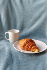 Fresh croissant on plate near tea in white cup on blue blanket in bed. Vertical shot. Cozy morning at home.