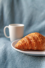 Tasty croissant on plate near coffee in white cup in bed. Vertical close up shot. Cozy morning at home.
