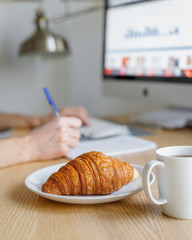 Fresh tasty croissant and a cup of coffee against the background of the workplace of a businessman, manager, freelancer, financial analyst. Coffee time at work concept. Remote work. Stay home concept.
