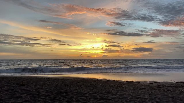 A slow motion shot tilting down from an epic sunset with incredible colors like a painting, onto rippling sand under the golden hour light on a beach in tropical Bali, Indonesia.