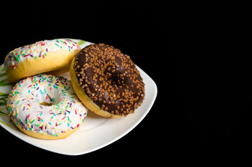 trend in food. chocolate and white icing donuts with different sprinkles and jam fillings on plate on black background with copy space