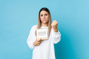 Young caucasian woman holding a 2020 calendar showing fist to camera, aggressive facial expression.