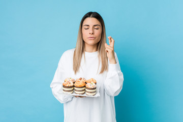 Young caucasian woman holding a sweets cake crossing fingers for having luck