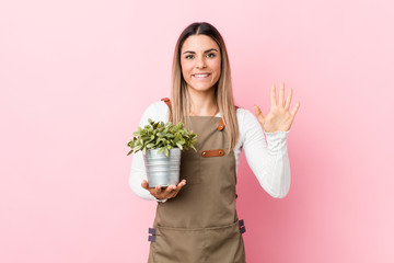 Young gardener woman holding a plant smiling cheerful showing number five with fingers.