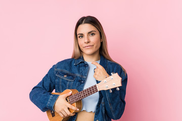 Fototapeta premium Young caucasian woman playing ukelele pointing with finger at you as if inviting come closer.