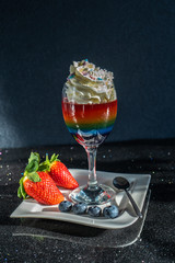 Colored rainbow jelly, layered jelly in a glass on a white plate with strawberries and blueberries vertical on a dark background