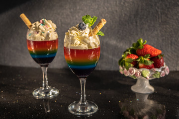 Colored rainbow jelly, layered jelly in a glass with whipped cream and blueberries on a dark background